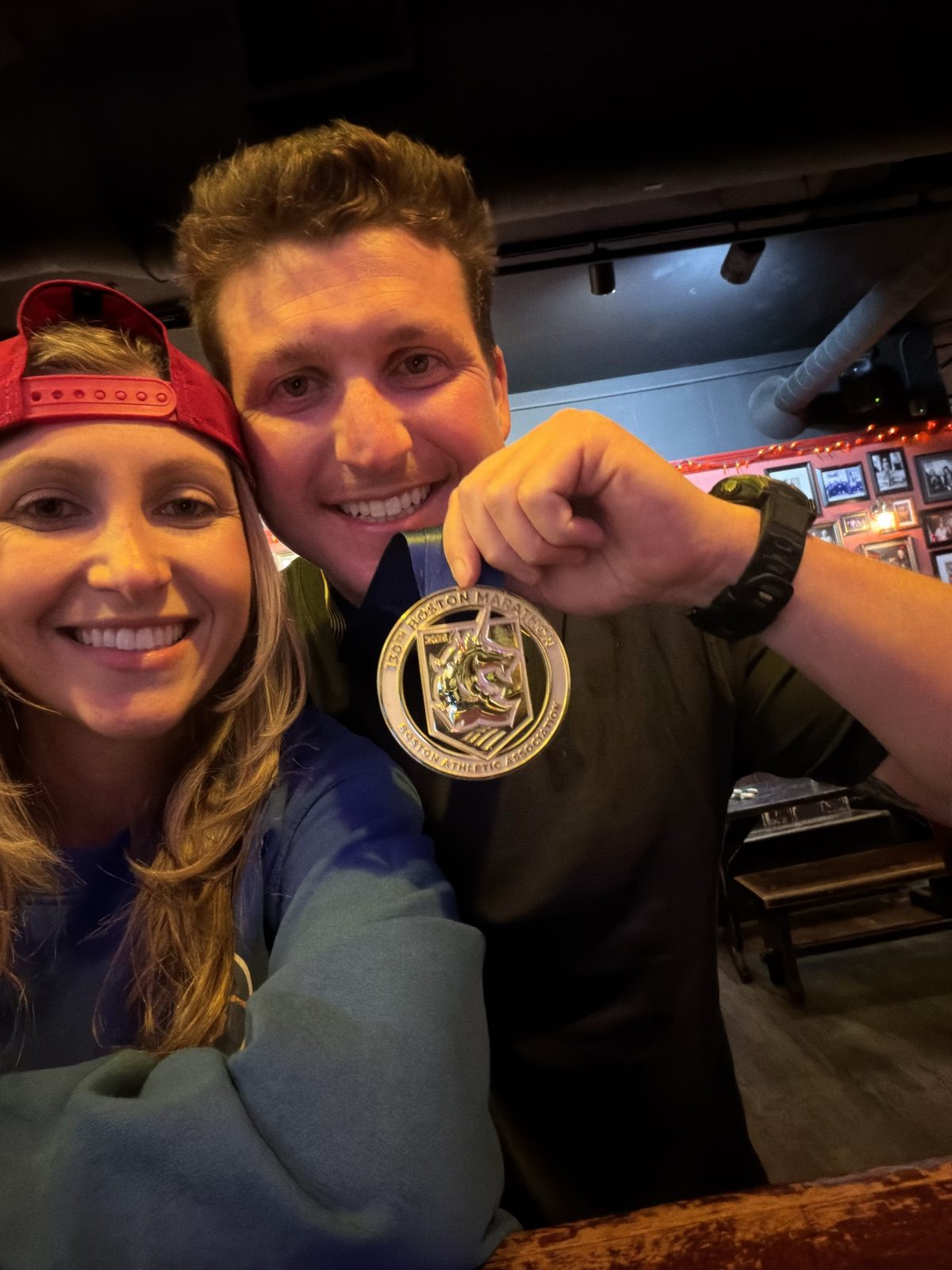 Andrew holding up his Boston Marathon medal with Jenna leaning in, celebrating at a bar