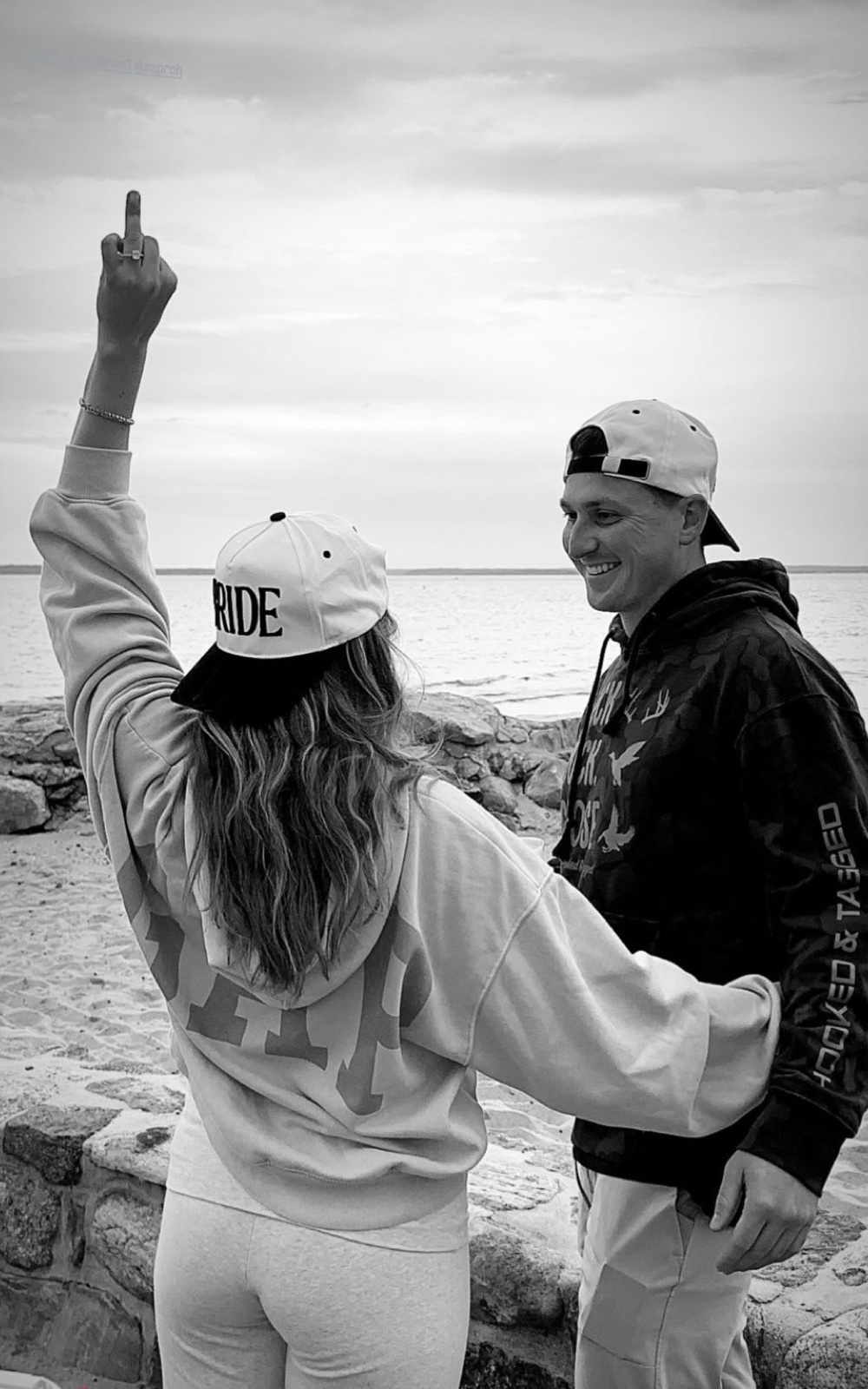 Jenna in a BRIDE hat pointing to the sky with her ring on, Andrew laughing beside her