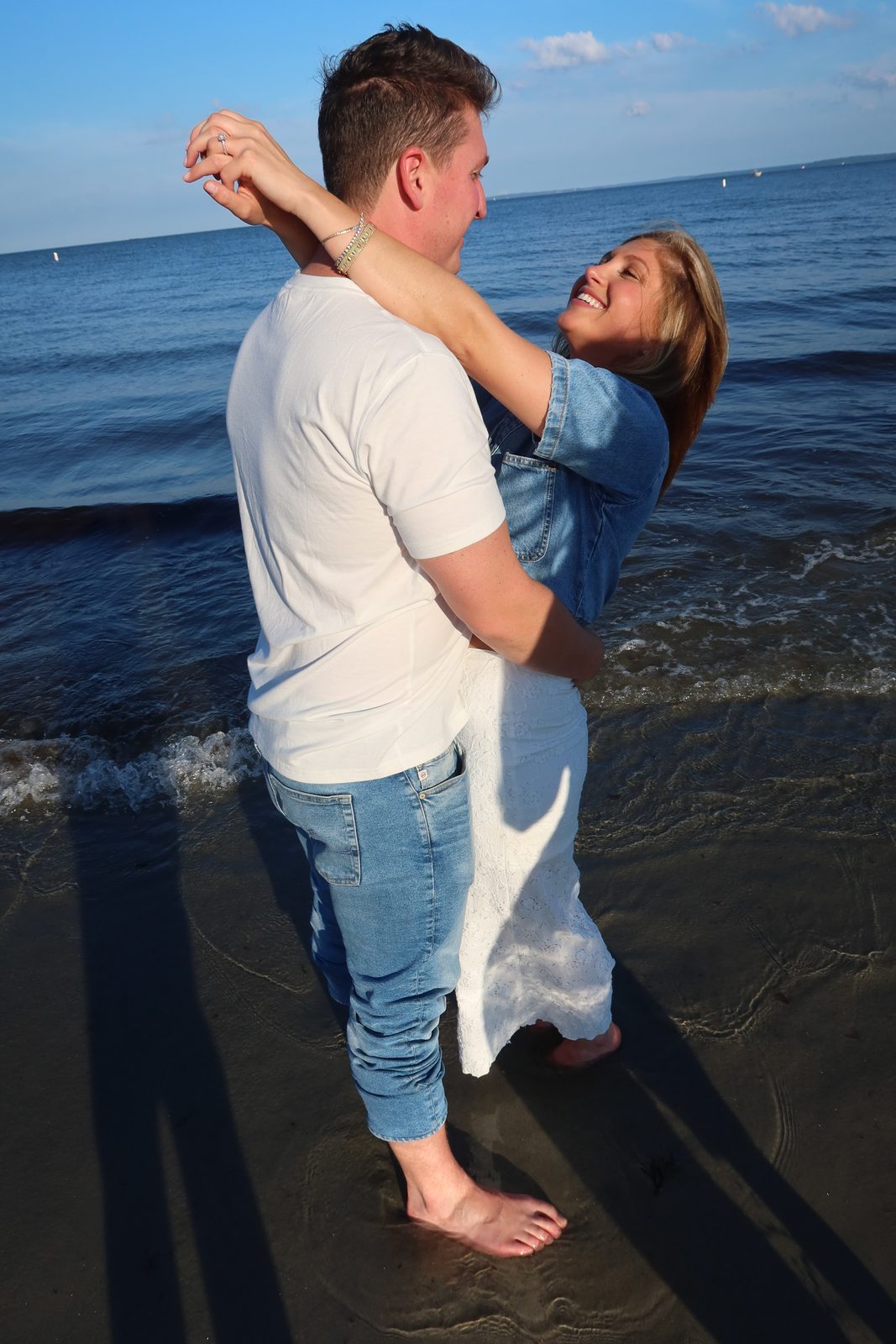 Jenna with her arms around Andrew's neck, both barefoot in the surf