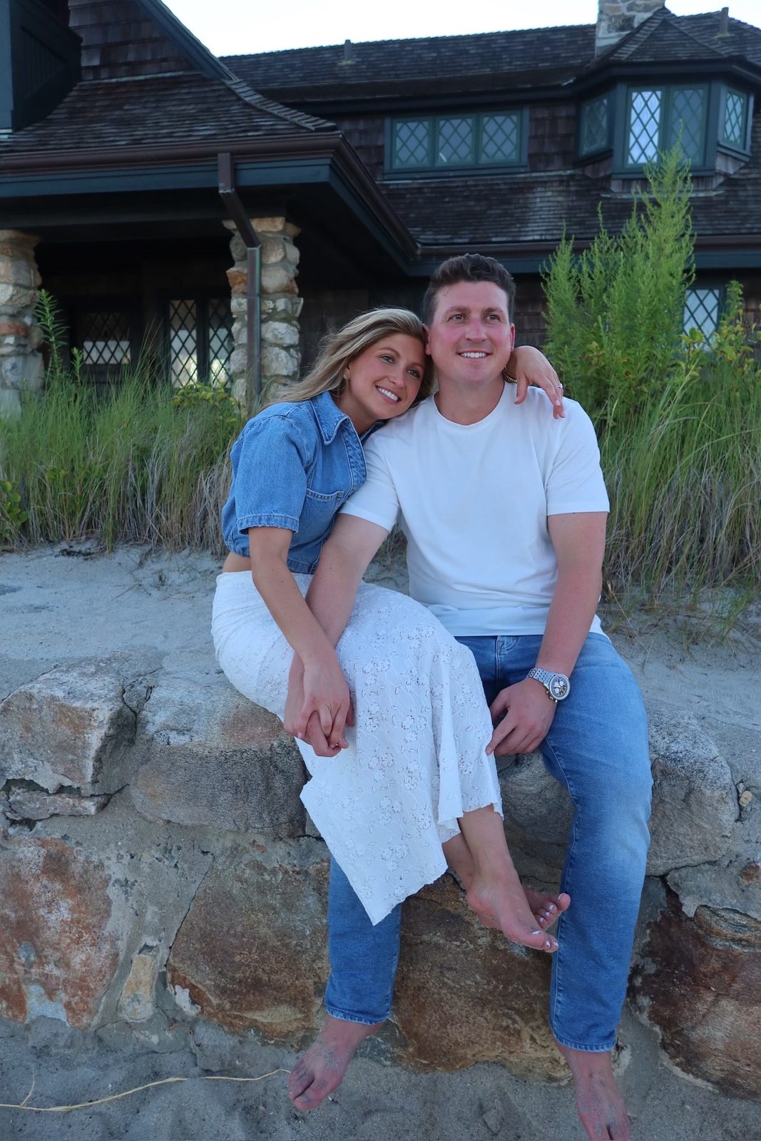 Andrew and Jenna sitting close together on a stone wall in front of a shingled beach house