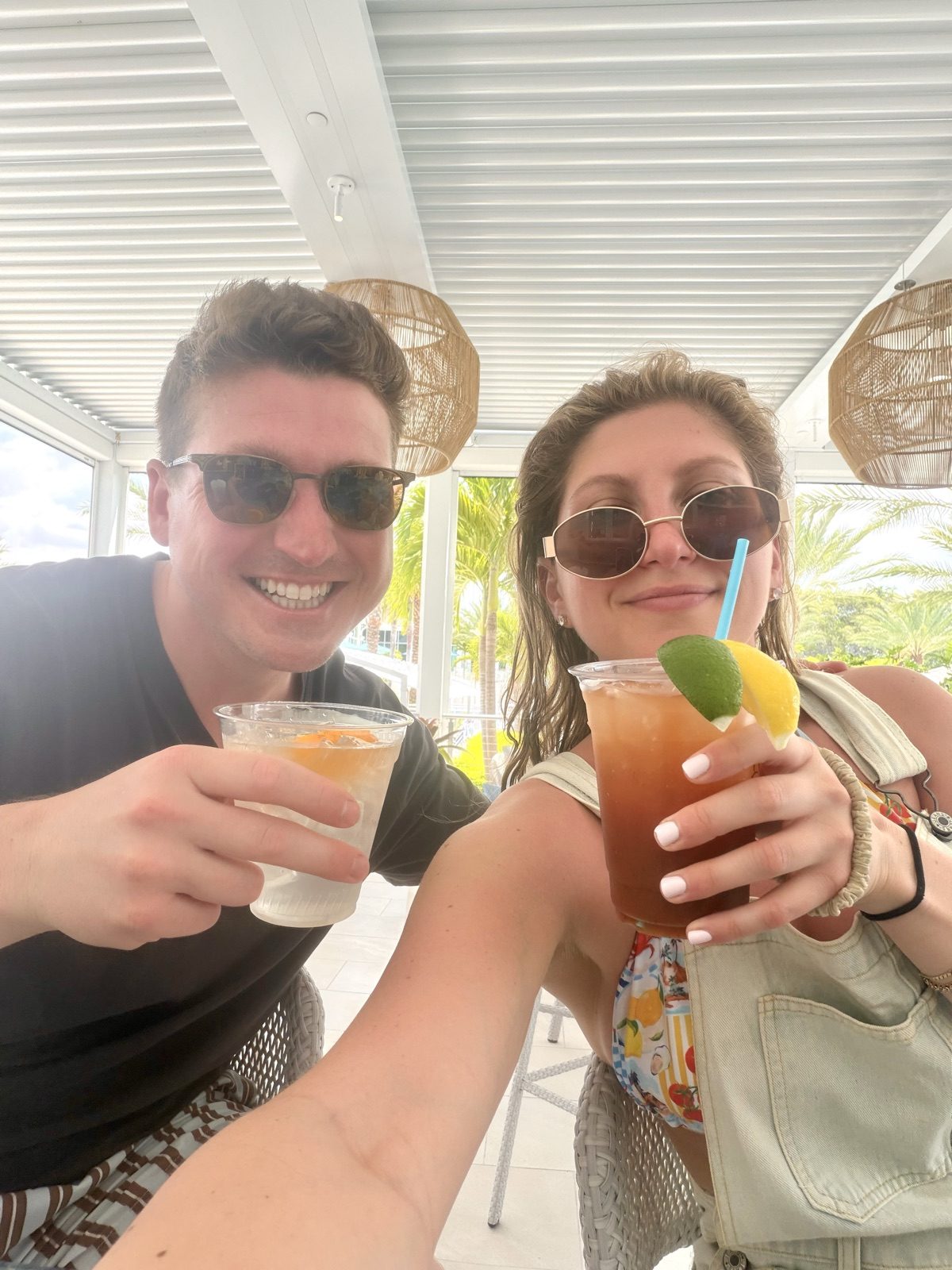 Selfie of Andrew and Jenna toasting drinks under a poolside pergola with palm trees behind