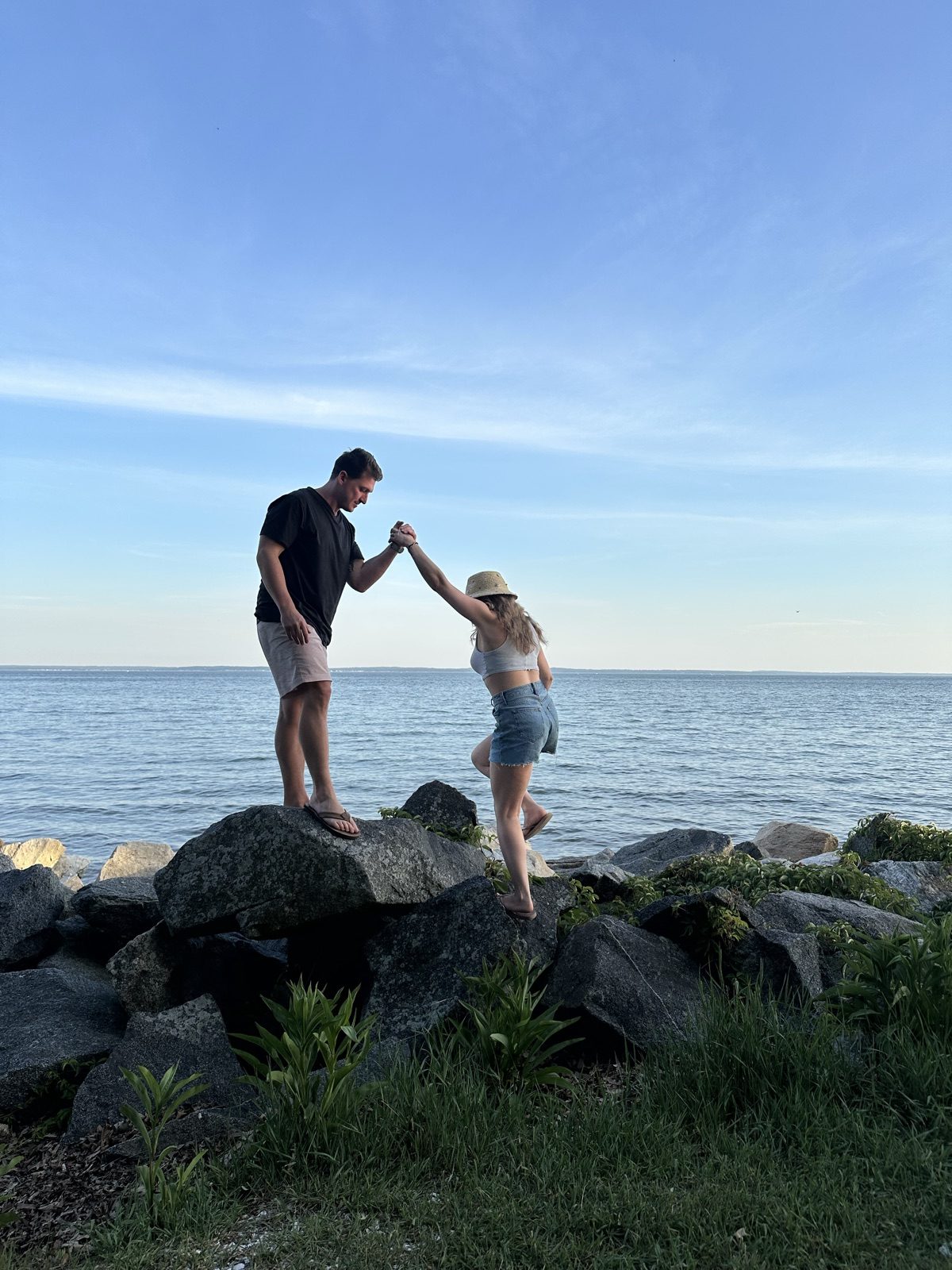 Andrew helping Jenna climb across shoreline rocks by the water